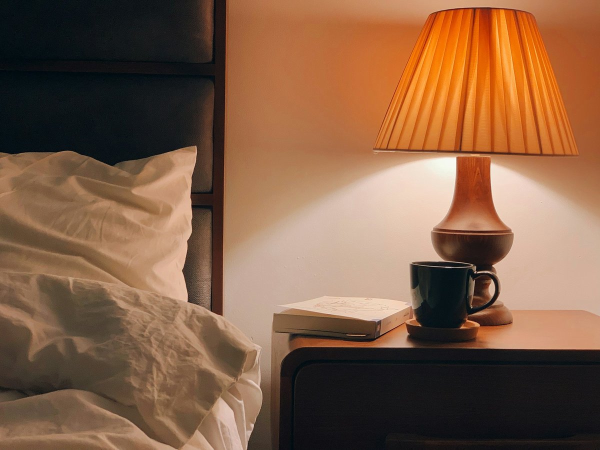 Lampe de chevet allumée près d'un lit, avec une tasse et un livre posés à côté